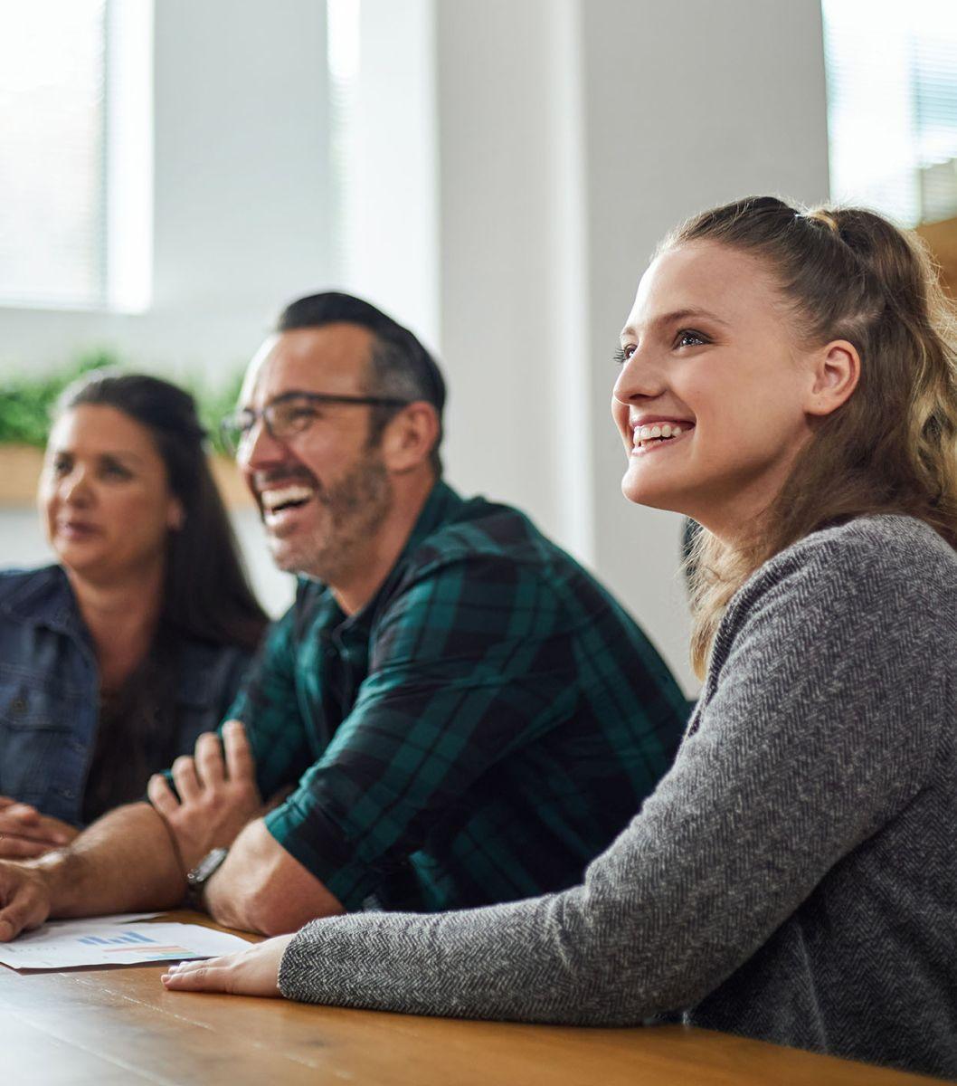 Three people sitting at wooden table in bright room all looking off camera in the same direction.