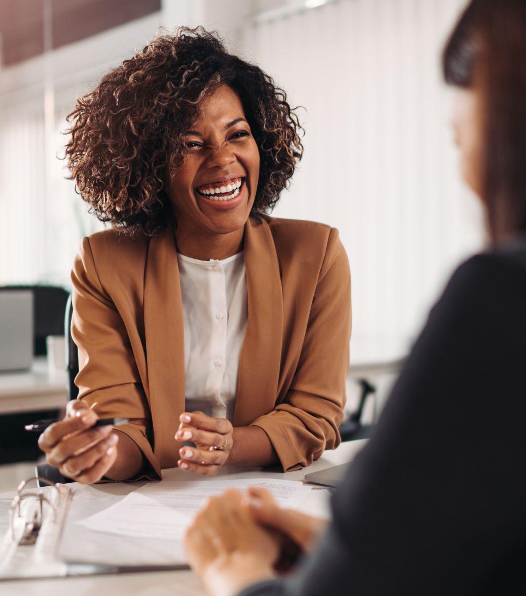 Woman wearing brown jacket and holding a pen, smiling as she looks at her client sitting across from her.