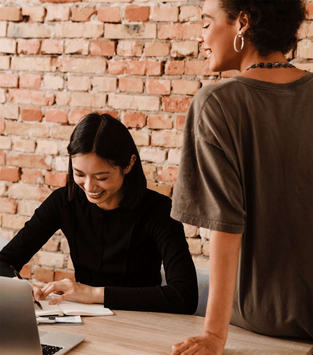 Women sitting at desk in black shirt, working. Another women standing nearby. Brick wall in the background.