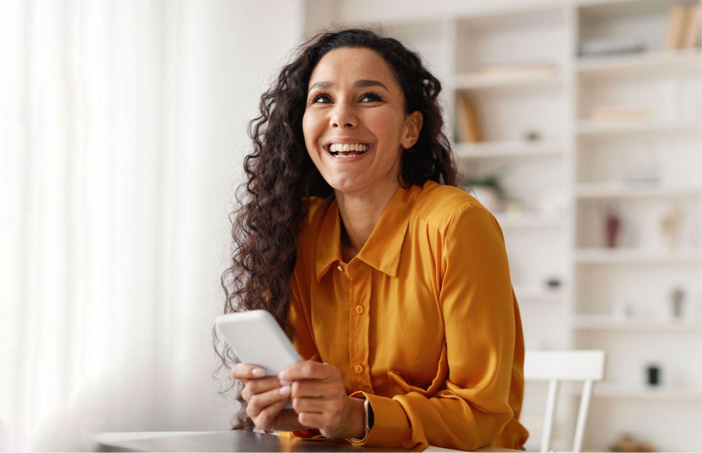 Smiling woman holding a smartphone, sitting at a desk in a bright room.